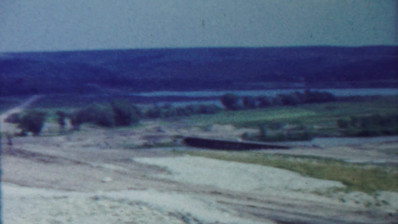 Wide rural landscape with dirt road, pale sky, low hills, and sparse trees.