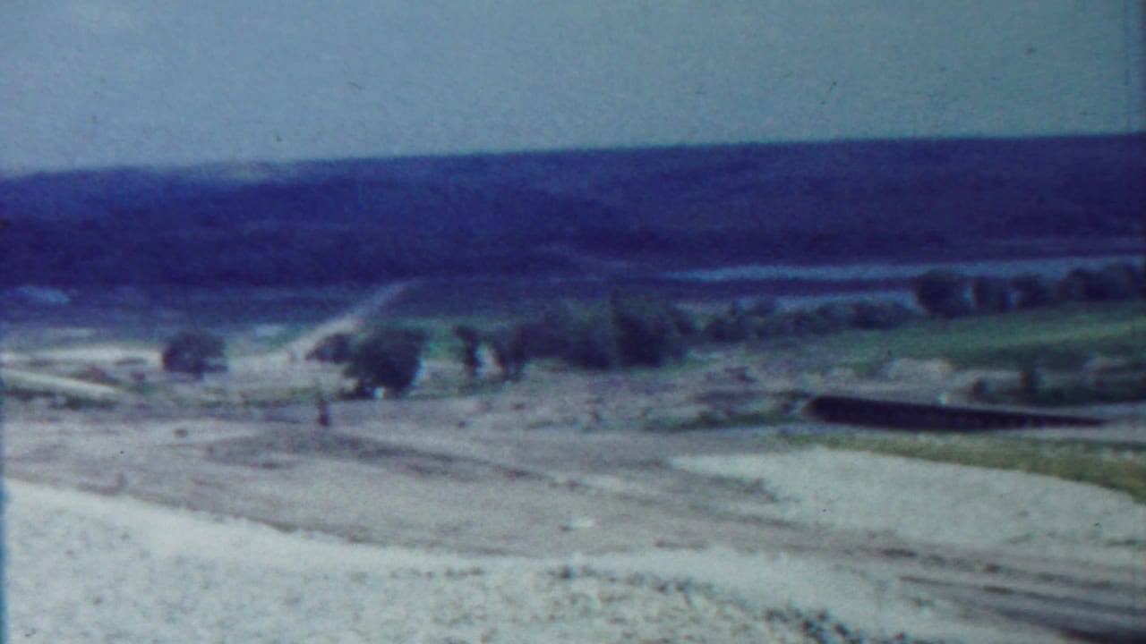 Dirt road crossing open terrain with low hills, trees, and distant water or lowland.