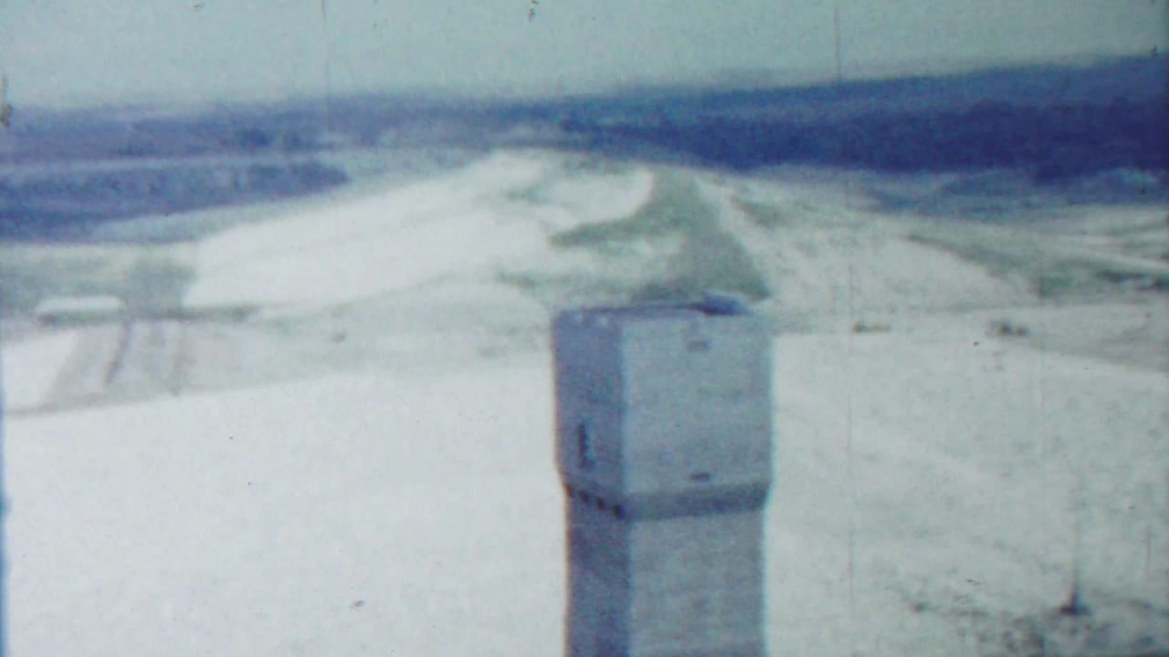 Concrete tower at lower center with sandy terrain and distant fields beyond.