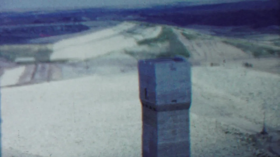 1959 Kansas 8mm view of a concrete tower with pale ground, fields, and a dark distant tree line.