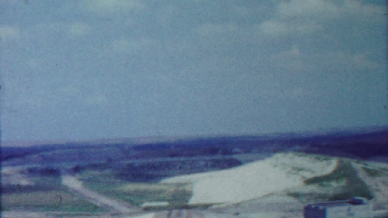 Wide view of dam embankment, road, fields, distant hills, and blue sky.
