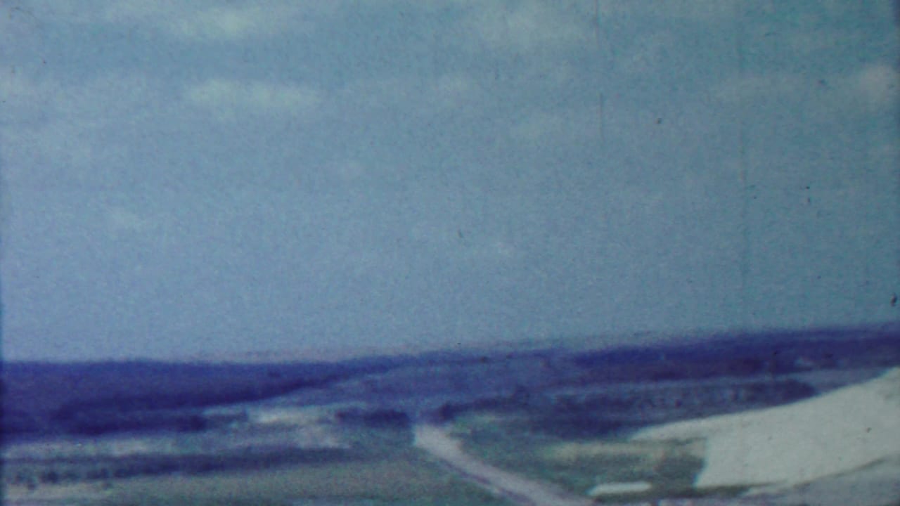 Distant dam landscape with pale embankment, fields, road, hills, and sky.
