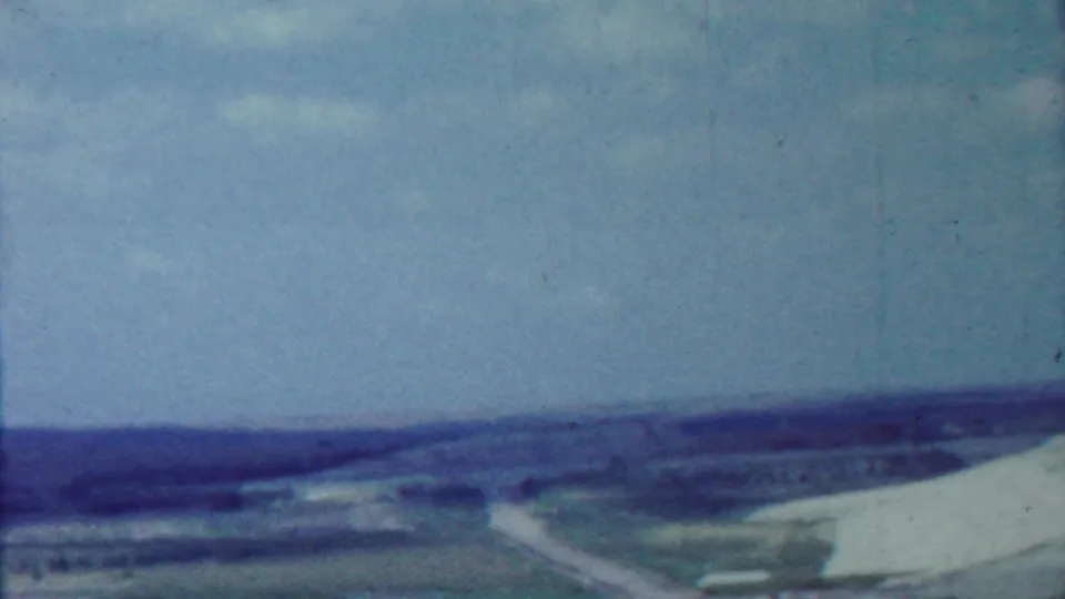 Visitor-shot 8mm frame of the Tuttle Creek Dam area, showing the pale dam slope, access road, fields, and low hills.