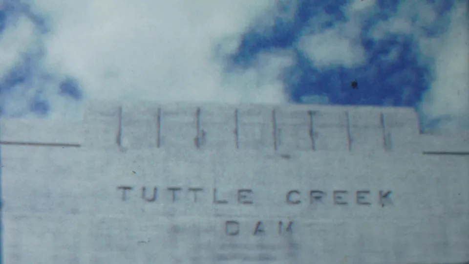 Close 8mm frame of the concrete structure labeled Tuttle Creek Dam, with trees above and visible film softness and scratches.