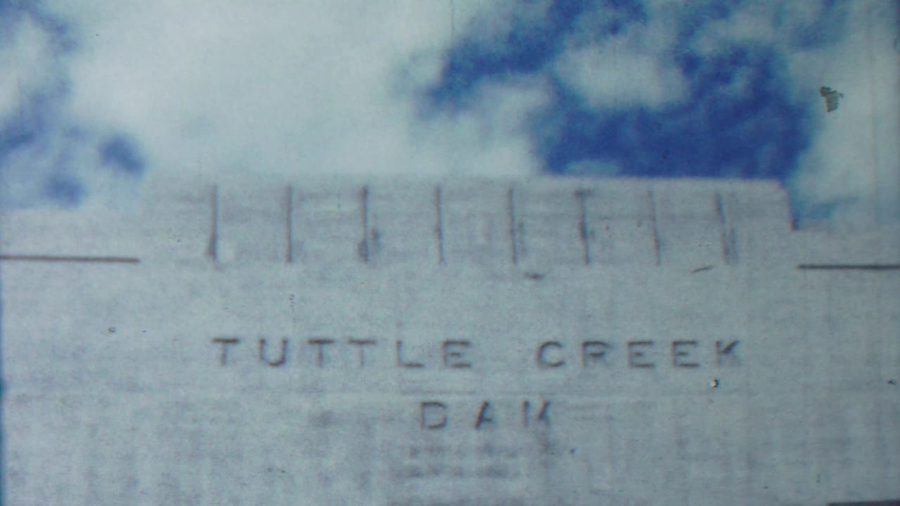 Concrete dam wall with faint Tuttle Creek Dam lettering beneath sky and trees.