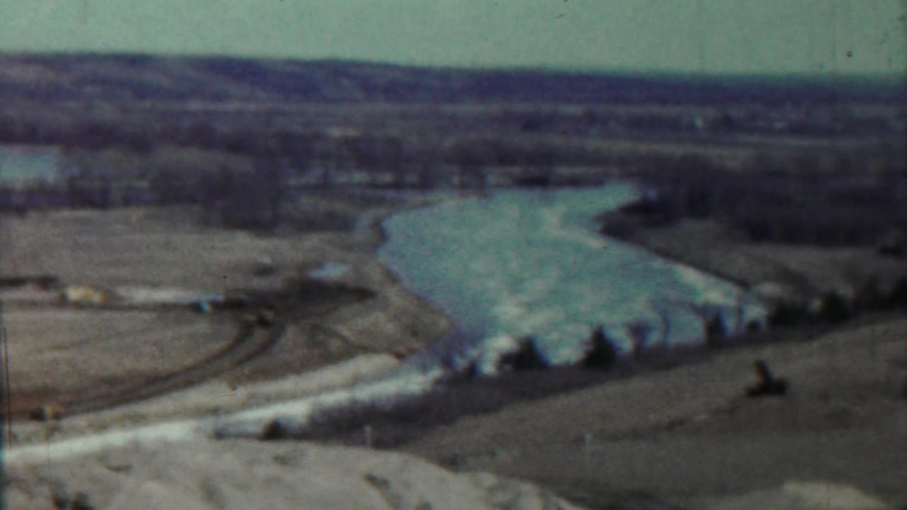 Wide lake view with curving water, low banks, tree line, and a small tan vertical form near shore.