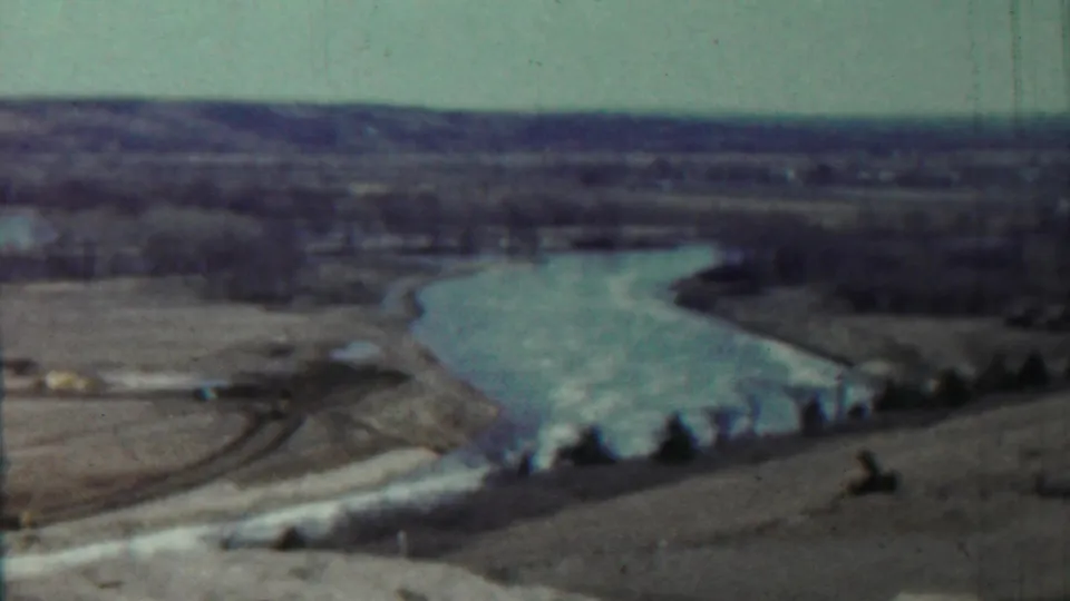 Faded 1959 8mm frame from Saint Francis, Kansas, showing a lake bend with pale shoreline, open fields, and sparse tree cover.