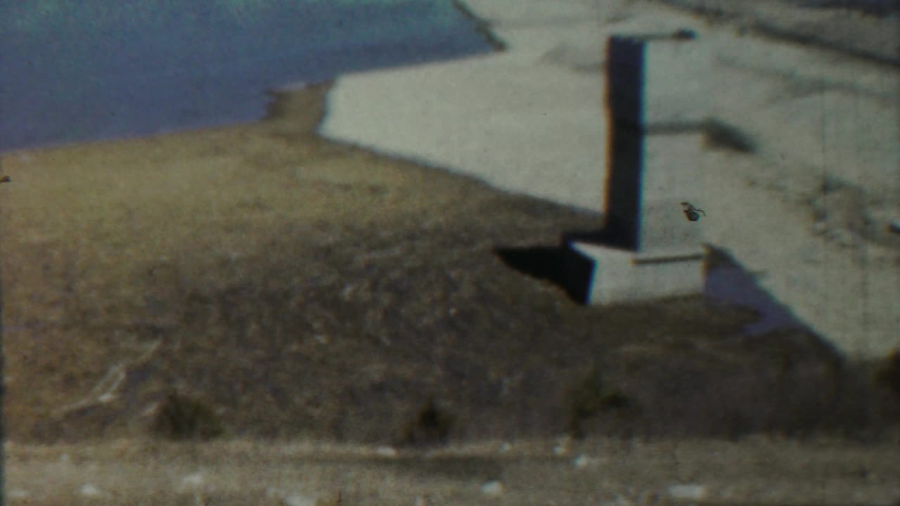 1959 Saint Francis, Kansas 8mm frame showing a pale column near a water edge, dry dirt, and sparse vegetation.