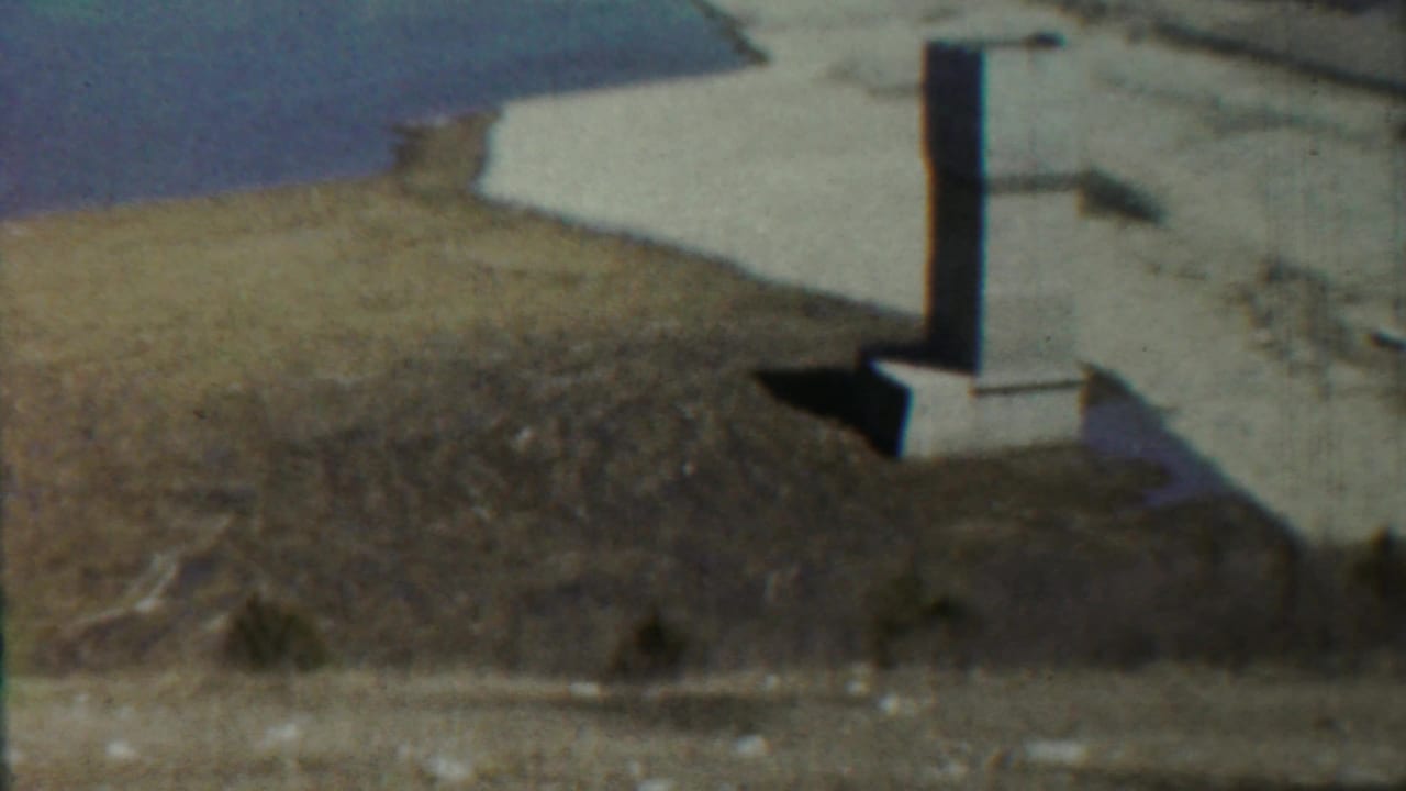 Column and base at the edge of dry land with water in the background.