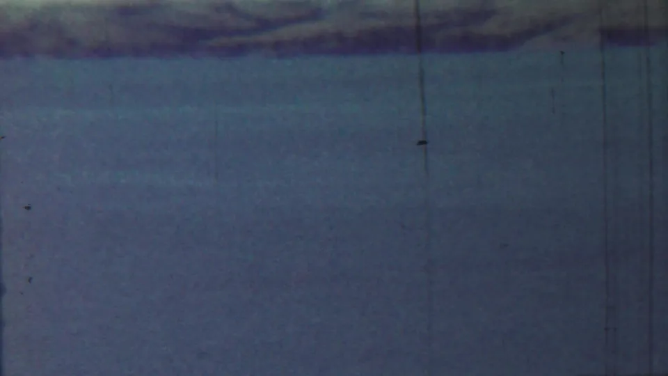 Archival 8mm view of hazy water and a dark riverbank, with dust specks and vertical scratches visible in the film scan.