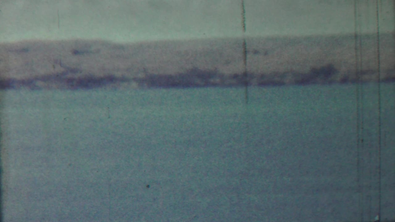Distant low shoreline across blue-green water beneath a pale sky, with visible film scratches.