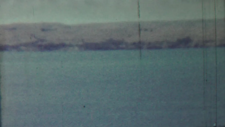 1959 Saint Francis, Kansas landscape frame with blue-green water, a far shoreline, pale sky, and vertical 8mm scratches.