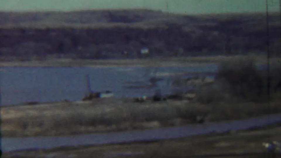 1959 rural Kansas home-movie frame showing a blue lake, dry grass, distant tree-covered hills, and a road or path in the foreground.