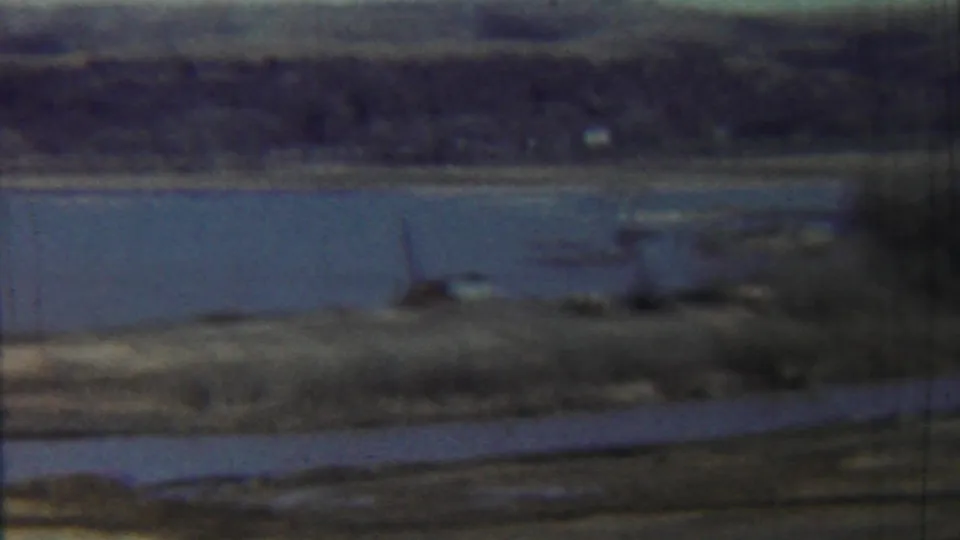 Saint Francis, Kansas 8mm frame showing lake water, a grassy bank, a foreground road, and small shoreline objects under a pale sky.