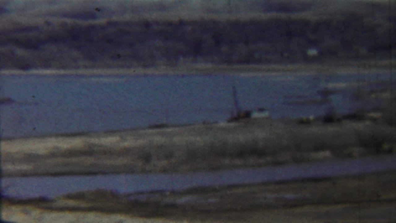 1959 Saint Francis, Kansas home-movie frame showing a broad waterway, distant shoreline, small boat, and low bridge or pier.