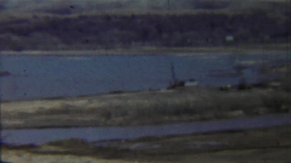 1959 Saint Francis, Kansas home-movie frame showing a broad waterway, distant shoreline, small boat, and low bridge or pier.