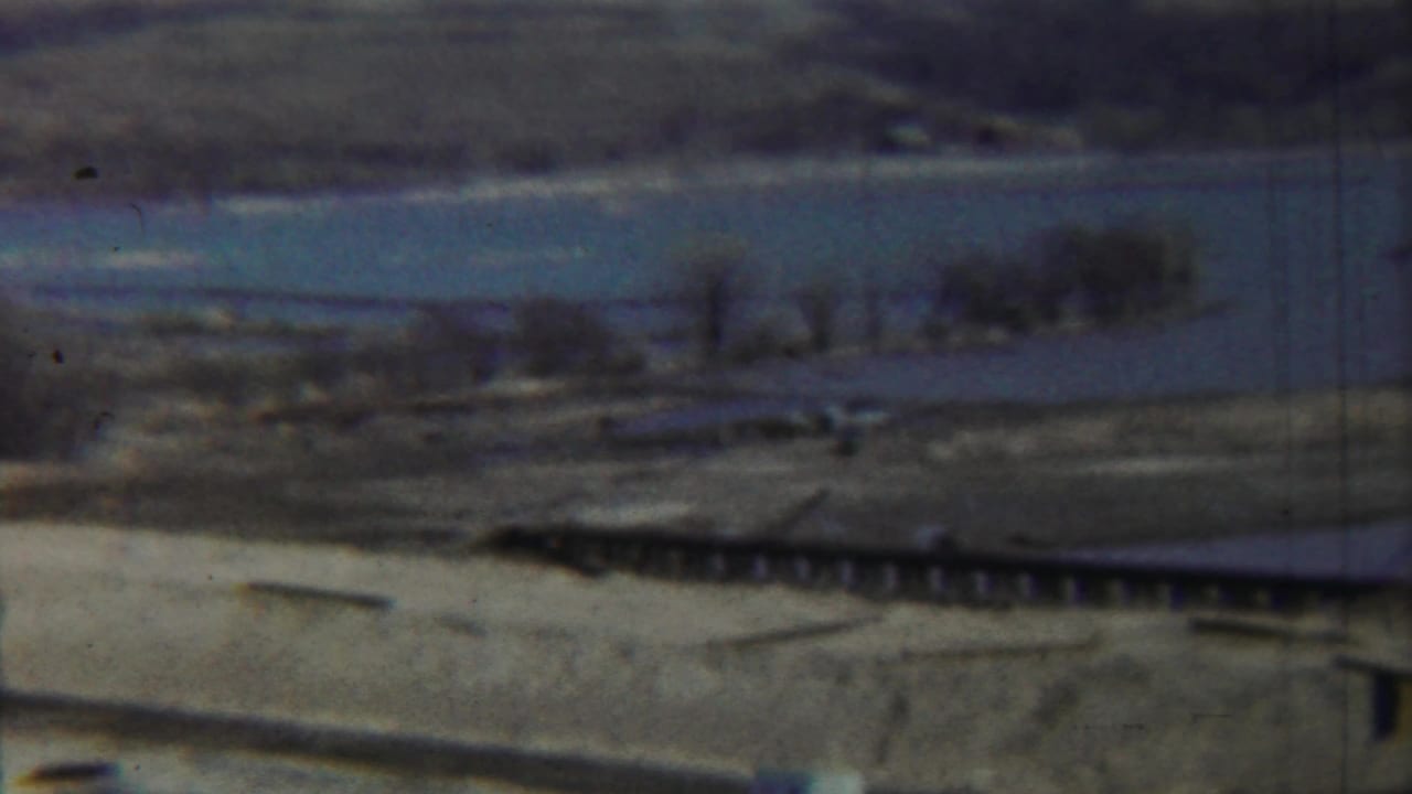 Hazy riverbed landscape with a low crossing, scrub, distant trees, and muted sky.
