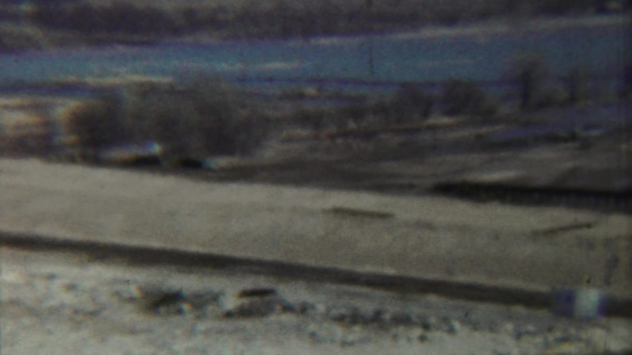 Wide arid landscape with a pale riverbed, low embankment, trees, and distant hills.