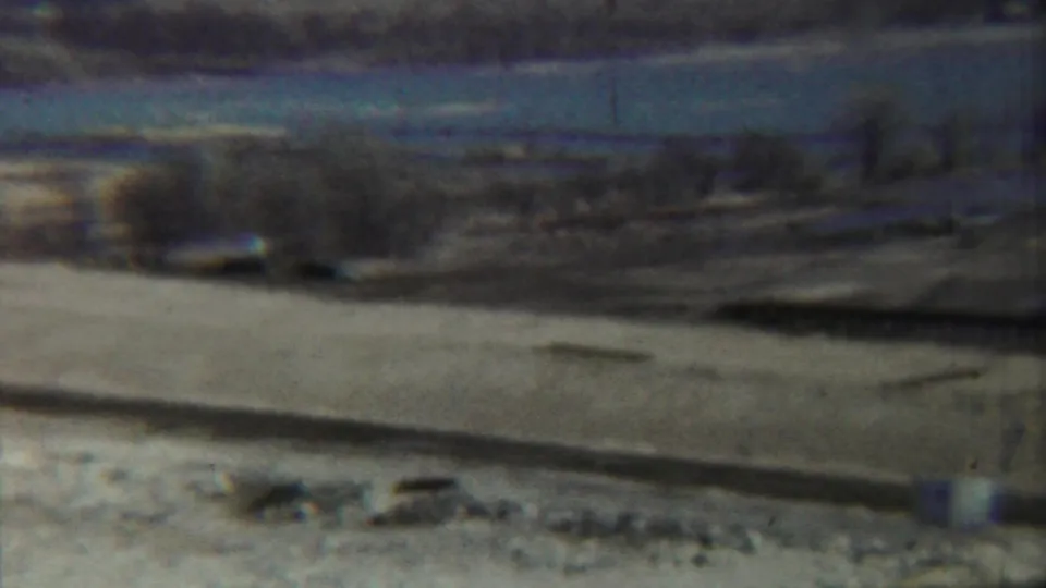 Archival 8mm frame from Saint Francis, Kansas, showing a washed, hazy view of a riverbed, low structure, sparse trees, and distant hills.