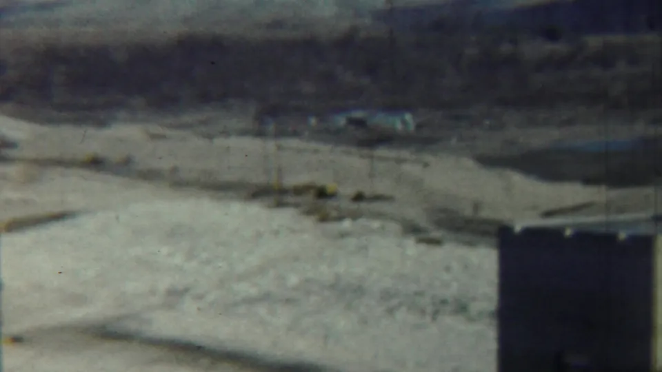 1959 rural Kansas frame showing a soft-focus riverbed or dry channel with a vertical support, scrub vegetation, and low hills beyond.