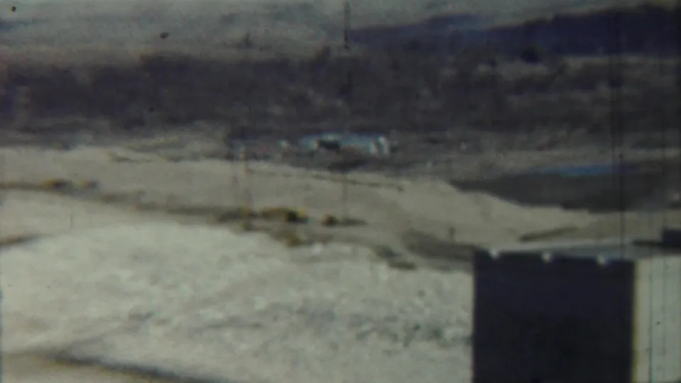 Archival home-movie frame near Saint Francis, Kansas, with a dark foreground support, pale riverbed, sparse brush, and hazy hills.
