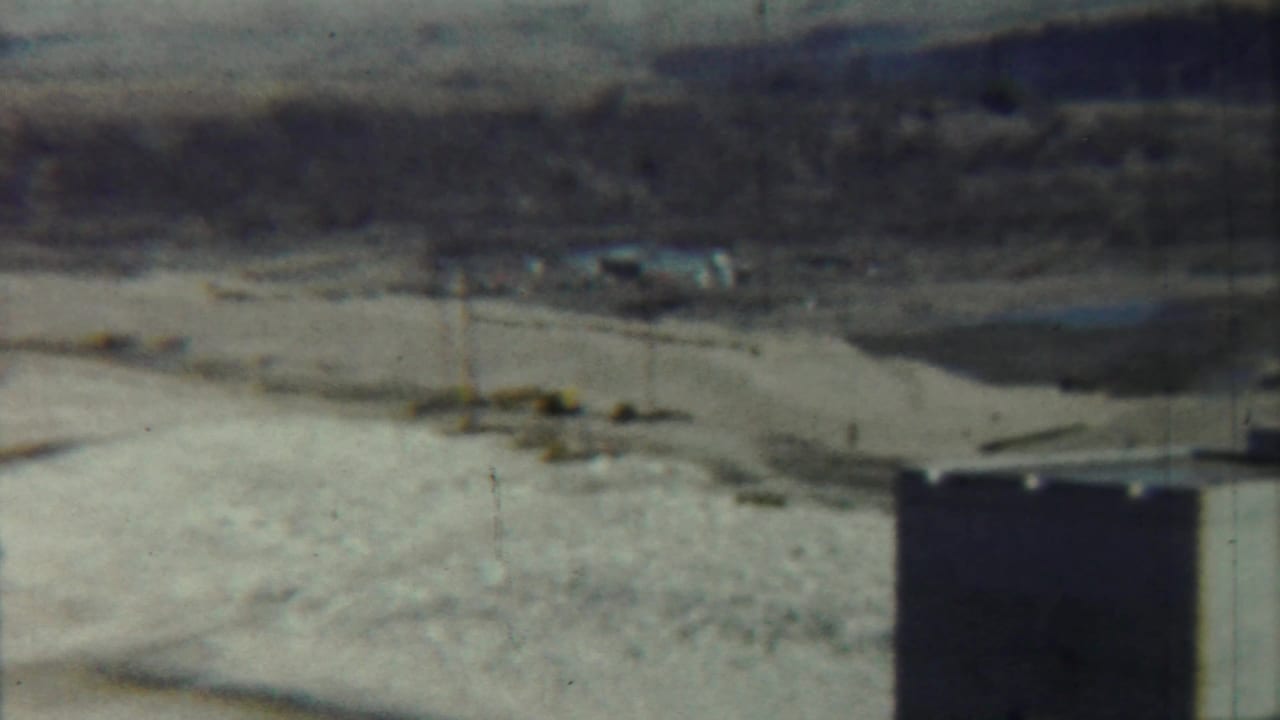 Soft 8mm view of a concrete tower edge beside a tan open field and distant structures.