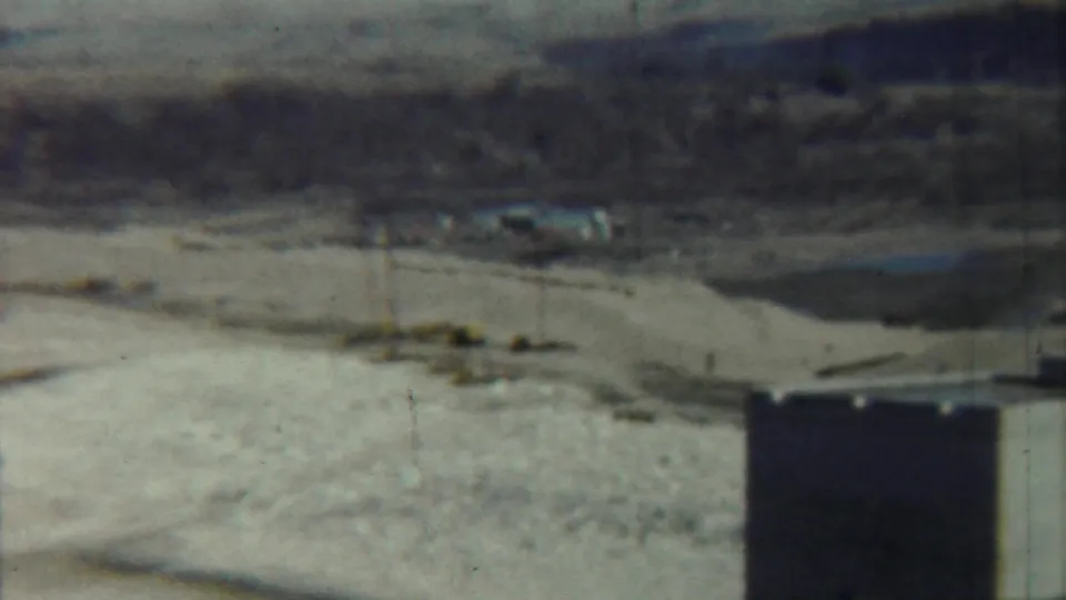1959 Saint Francis, Kansas 8mm frame showing a white concrete tower edge, open tan field, utility poles, and low distant buildings.
