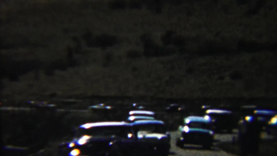 Nighttime Saint Francis, Kansas parking area with rows of classic cars, headlight glare, and indistinct people in shadow.