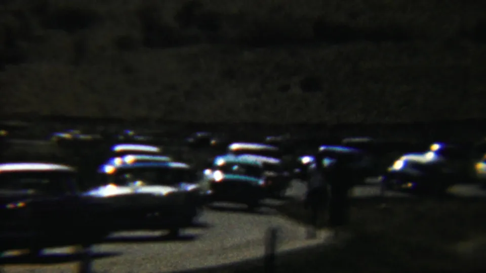 1959 home-movie frame of a busy nighttime parking area in Saint Francis, Kansas, with classic cars and shadowed figures.