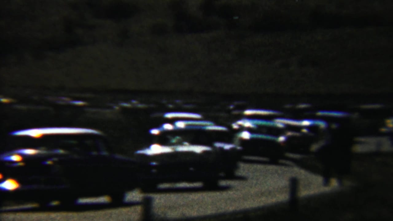 Dark classic cars lined up on a winding road at night with headlights glowing.