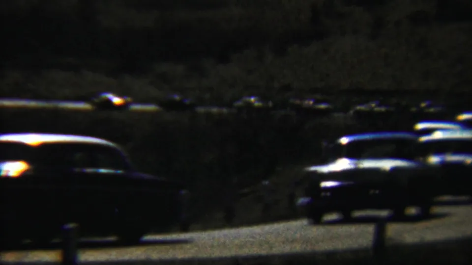 Low-light 1959 home-movie frame of classic cars rounding a road curve, with bright glare across windshields and roofs.