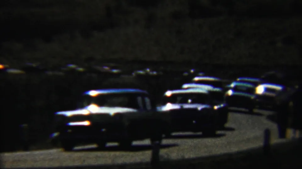 Saint Francis, Kansas 8mm frame showing late-1950s cars packed along a dim road, with indistinct figures near the vehicles.