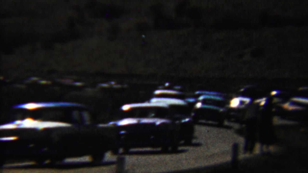 Low-light 1959 Saint Francis, Kansas home-movie frame showing classic cars on a road with people standing nearby.
