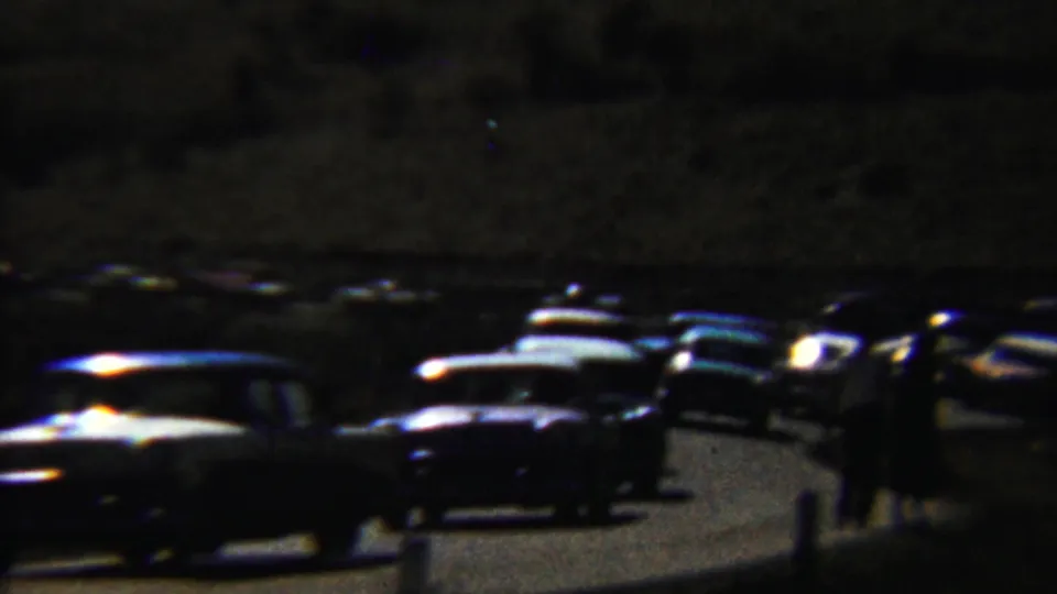 Low-light 1959 Saint Francis, Kansas home-movie frame showing classic cars on a road with people standing nearby.