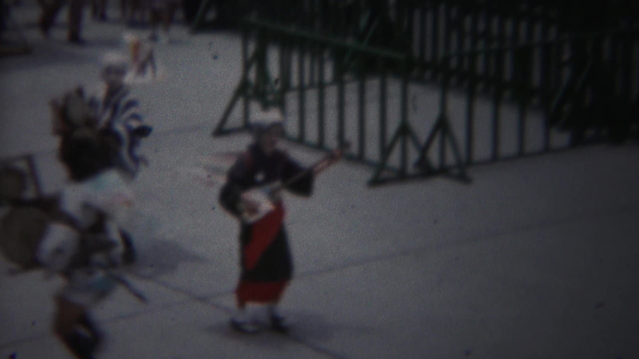 Costumed performers with instruments move beside green metal barricades on a paved street.