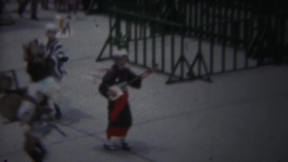 1954 Yokohama home-movie frame showing costumed street performers with instruments near green crowd barriers.