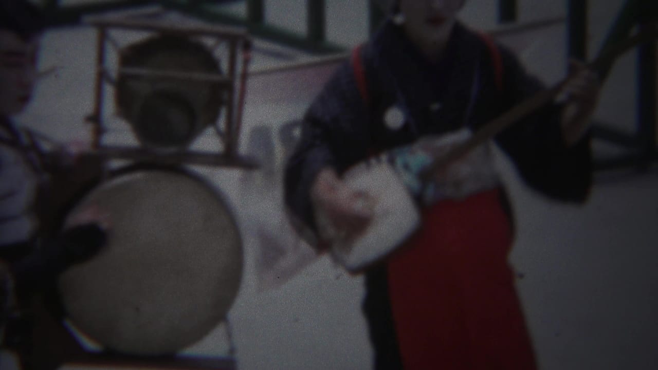 1954 Yokohama frame with performers in dark kimono and red hakama near a large drum and shamisen-like instrument.