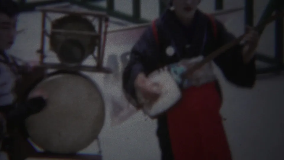 1954 Yokohama frame with performers in dark kimono and red hakama near a large drum and shamisen-like instrument.