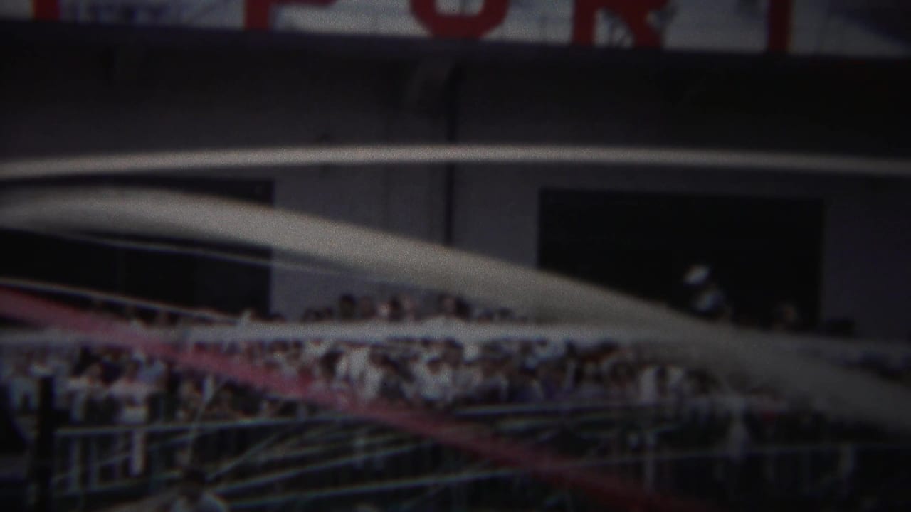 Crowd behind railings near parked 1950s cars and a large building.