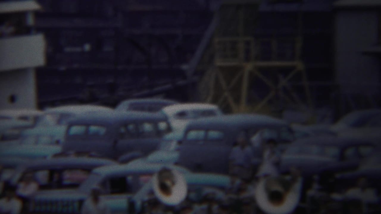 Yokohama 1954 frame showing classic parked cars with brass band members in the foreground and scaffold-like structures behind them.