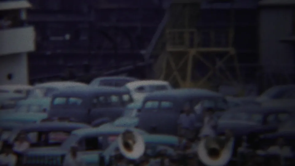 Yokohama 1954 frame showing classic parked cars with brass band members in the foreground and scaffold-like structures behind them.