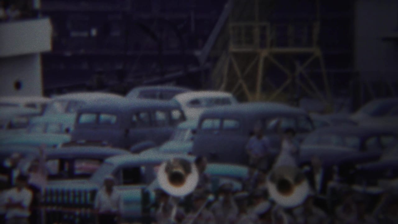 Uniformed brass band plays near parked 1950s cars as spectators gather at a port.