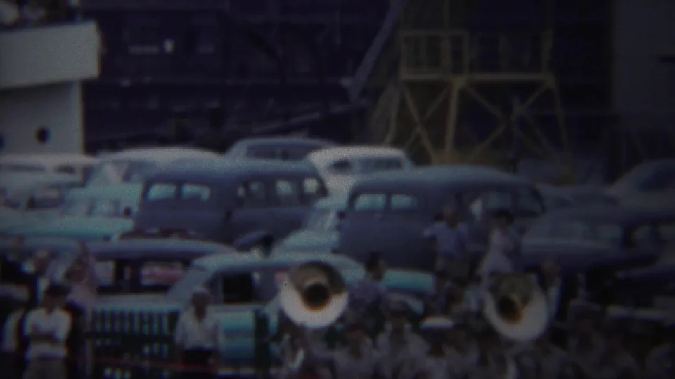 Yokohama 1954 frame showing brass players and a crowd among parked mid-century cars near an army port structure.
