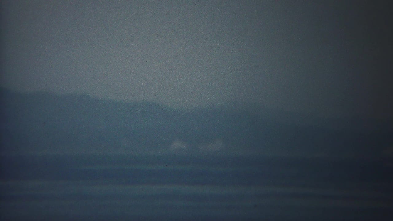 A 1954 Yokohama waterfront frame shows blue-gray water, shadowy hills, and faint pale shoreline details through haze.