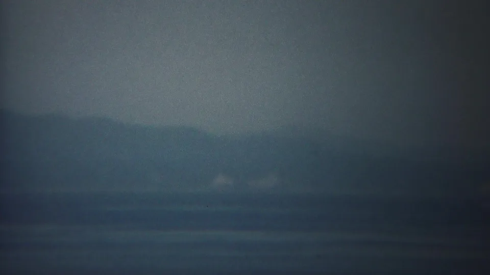 A 1954 Yokohama waterfront frame shows blue-gray water, shadowy hills, and faint pale shoreline details through haze.