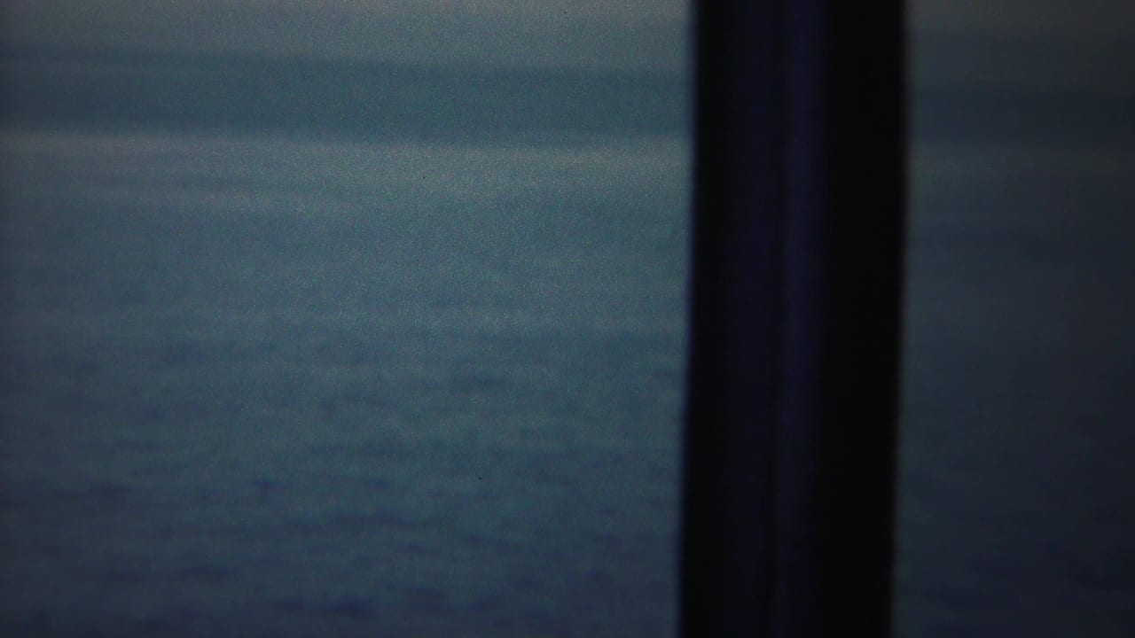 A 1954 shipboard home-movie frame looks through dark rail posts toward hazy blue water.