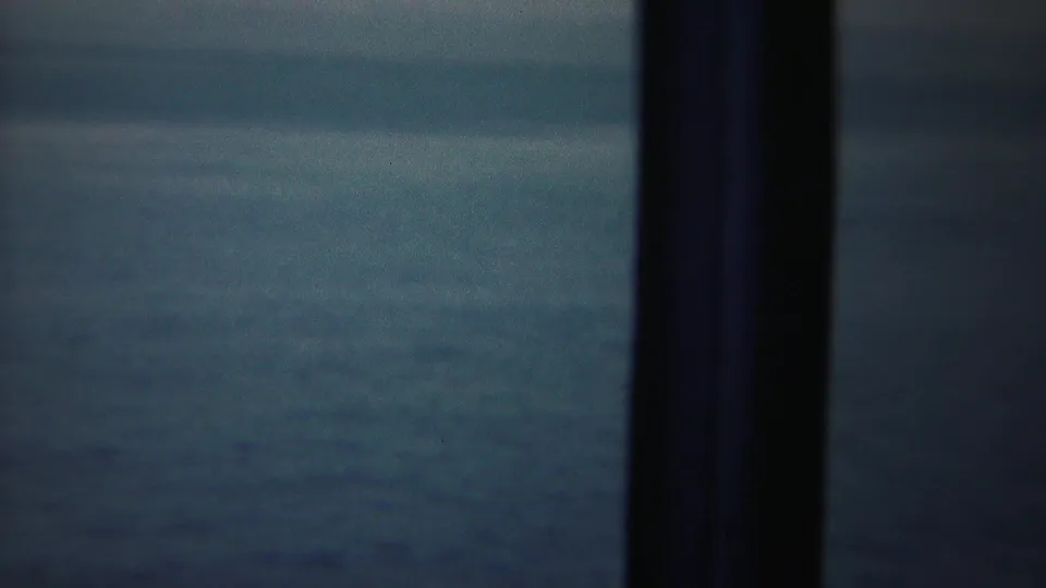 A 1954 shipboard home-movie frame looks through dark rail posts toward hazy blue water.