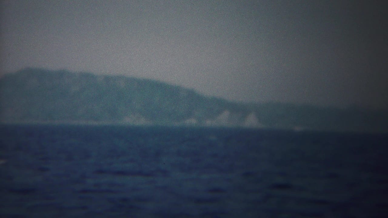 End frame of the 1954 8mm shoreline view, with blue sea, hazy wooded land, and pale coastal breaks in the distance.