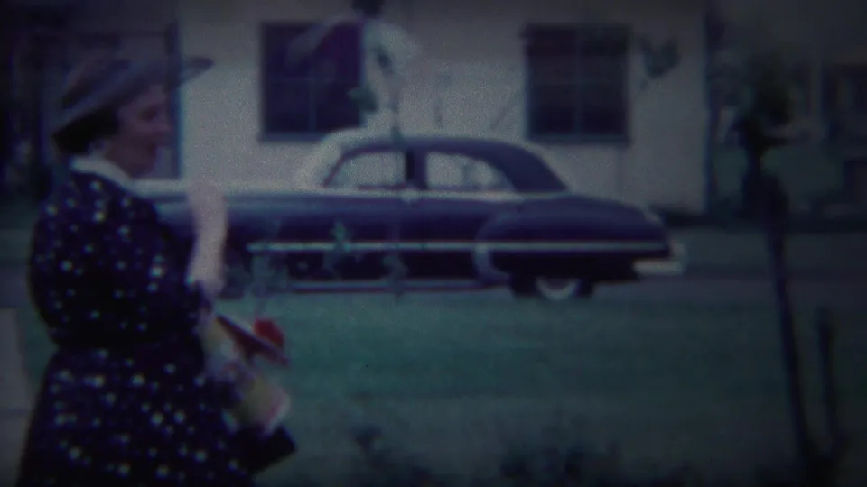A woman in 1950s attire walks outdoors in South Bend, Indiana, with a dark rounded sedan parked along the residential street behind her.