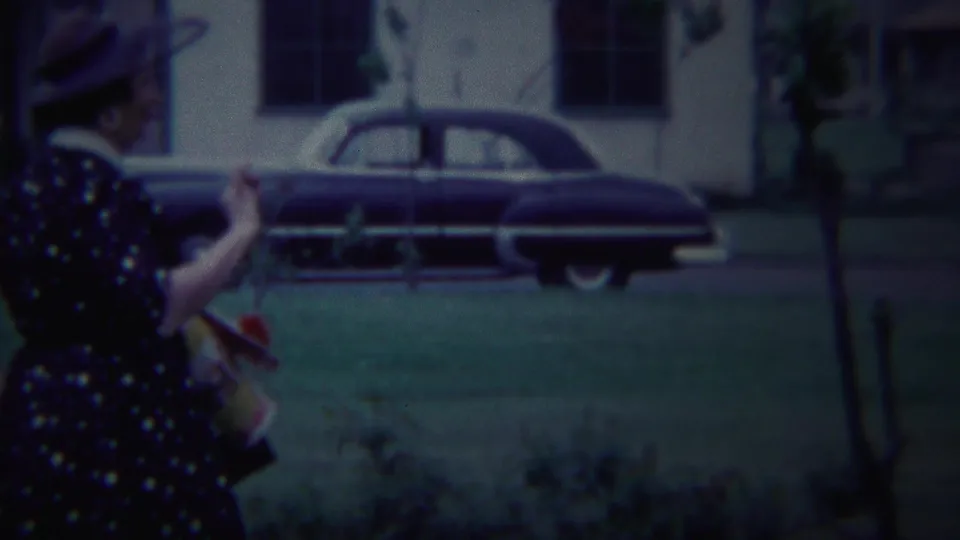 The walking woman is partly cropped at the left edge while a dark 1950s sedan, lawn, trees, and a residential building fill the background.